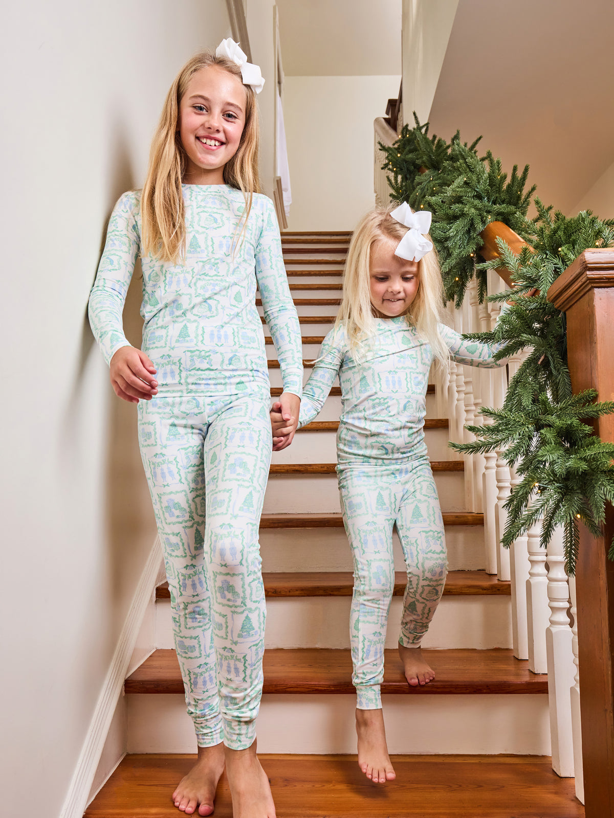Two young girls walking down wooden stairs indoors, wearing matching long-sleeve and leggings pajama sets with a light mint green and white geometric pattern. The older girl wears a white bow in her hair, and the stairs are decorated with a green garland.