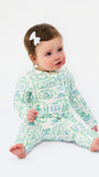 A baby sitting on a white studio background wearing a long-sleeve footed pajama set with a green and blue abstract pattern on a white base, accessorized with a small white bow in the hair.