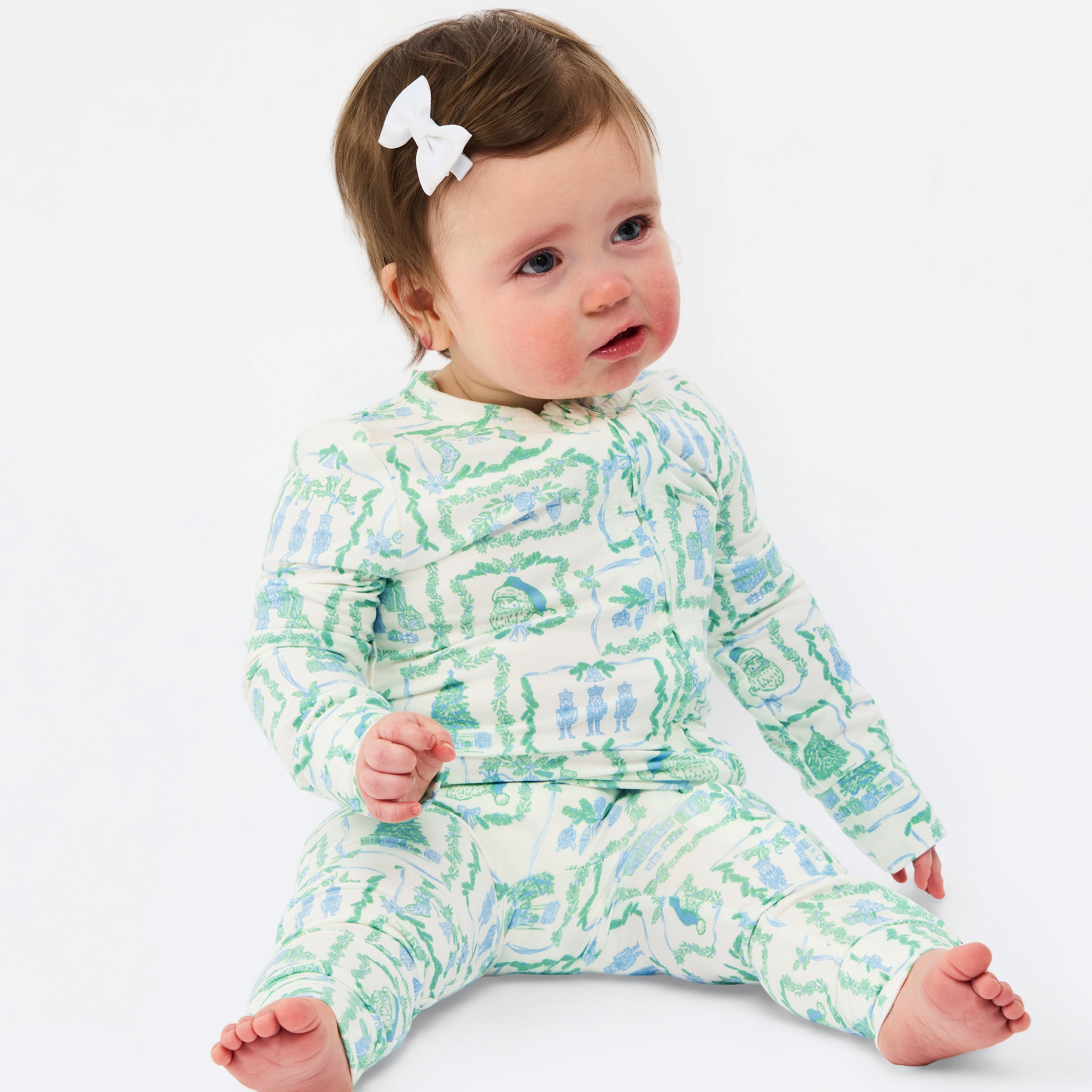 A baby sitting on a white studio background wearing a long-sleeve footed pajama set with a green and blue abstract pattern on a white base, accessorized with a small white bow in the hair.