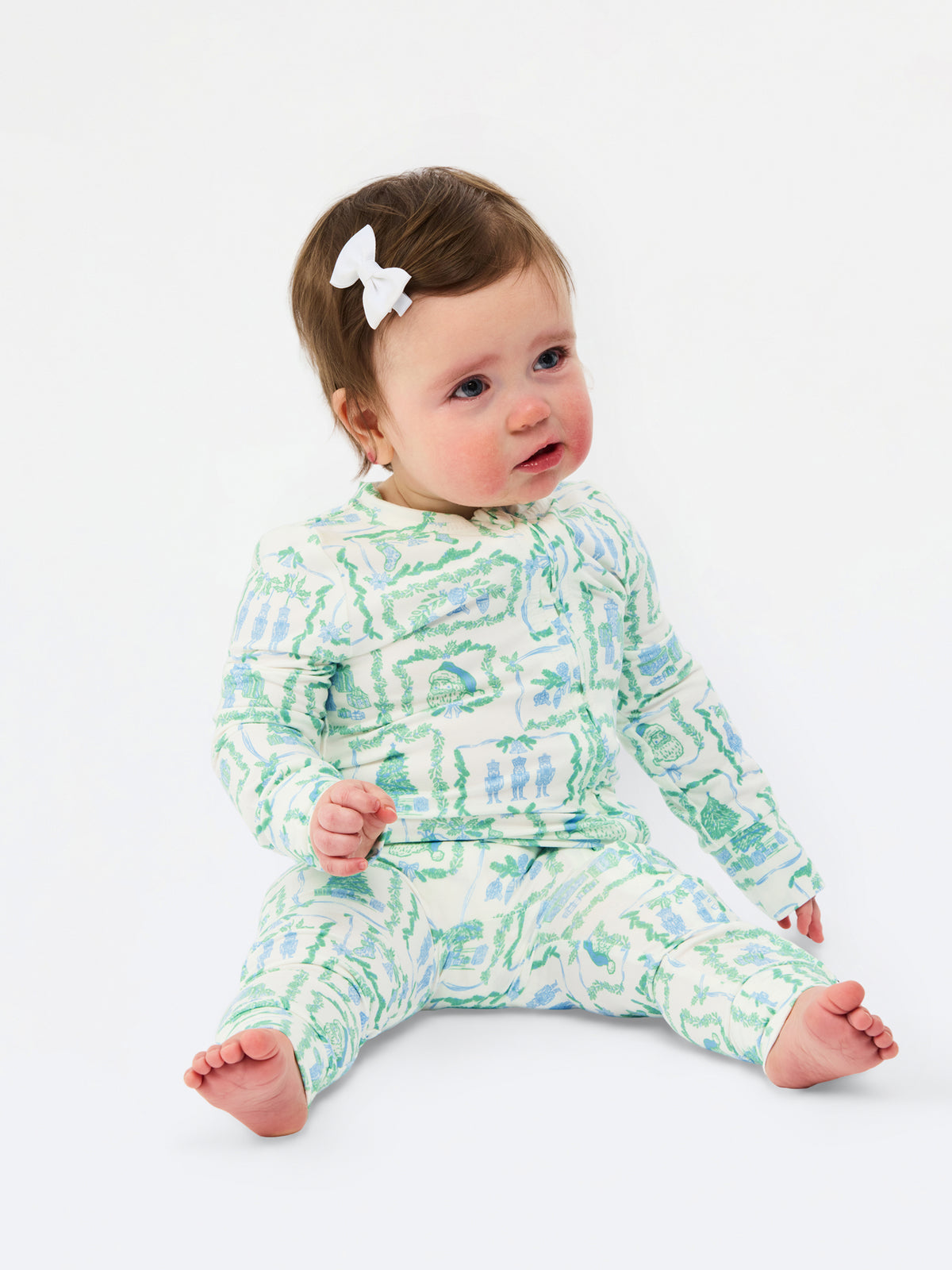 A baby sitting on a white studio background wearing a long-sleeve footed pajama set with a green and blue abstract pattern on a white base, accessorized with a small white bow in the hair.