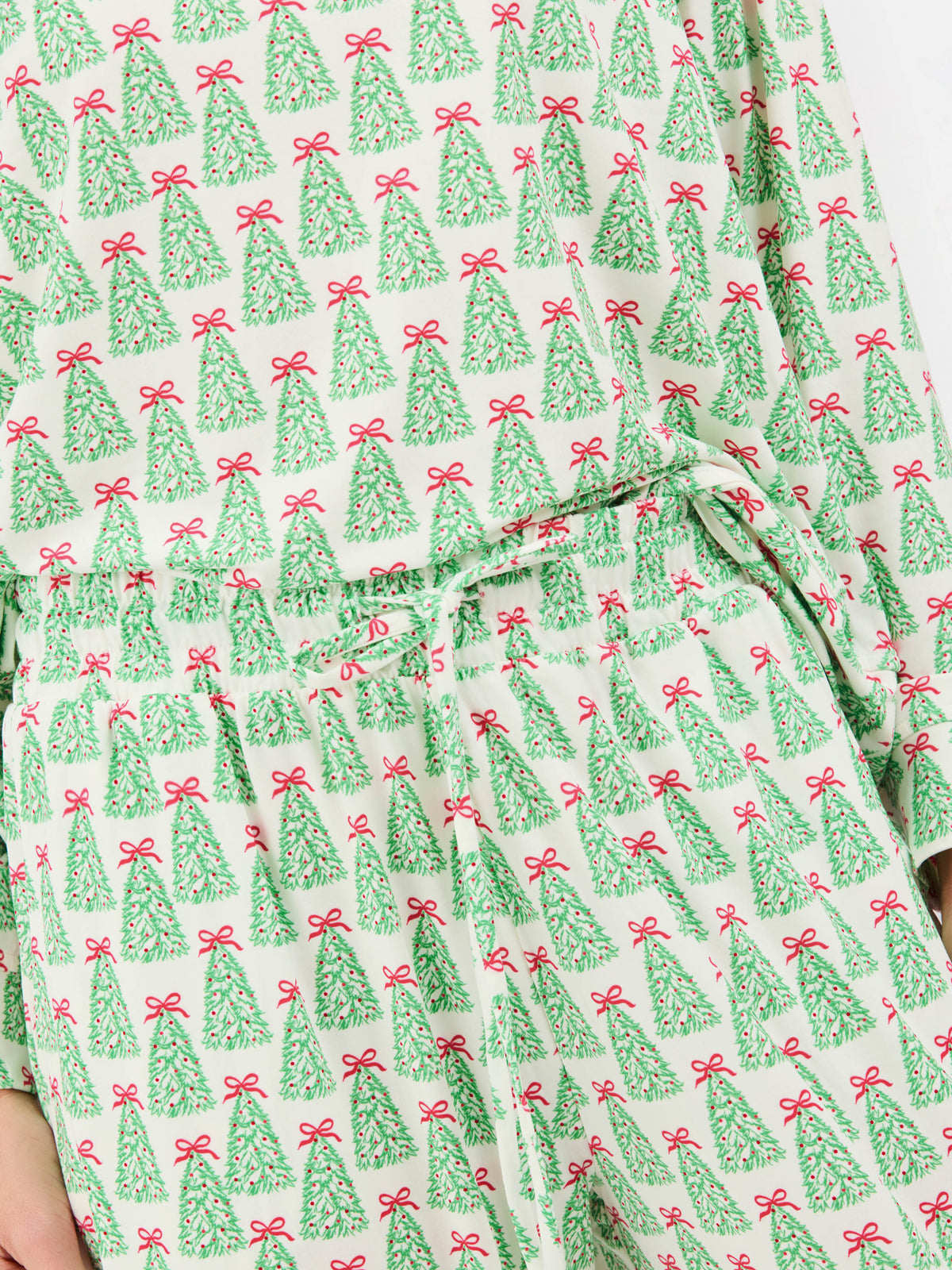 A close-up of a light-colored fabric garment with an all-over Christmas tree print. The pattern features green pine trees with red bows, and a matching drawstring waistband is visible at the center.