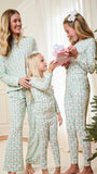 Three women of different ages wearing matching pajamas with a green pine tree pattern, standing in a living room next to a Christmas tree and receiving a gift.