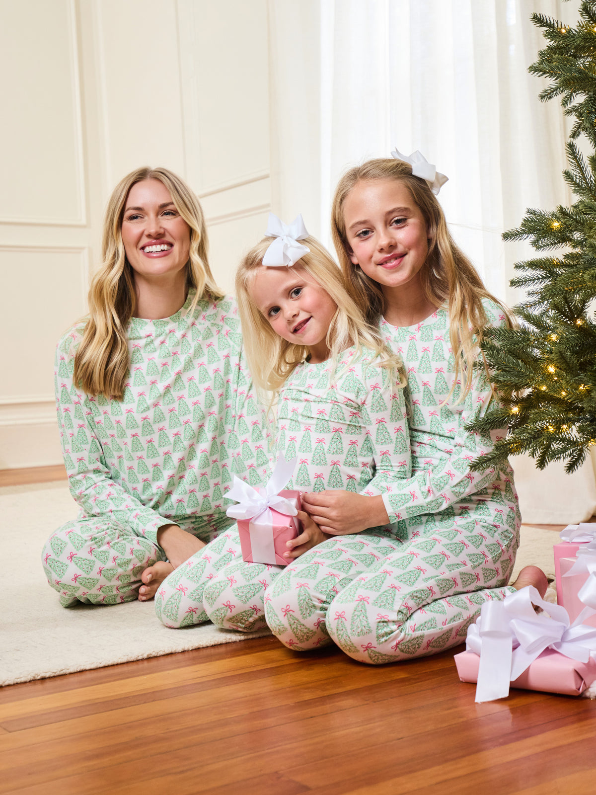 Three women wearing matching light green and pink Christmas tree print pajamas sit together on a wooden floor in front of a decorated Christmas tree, holding wrapped presents.