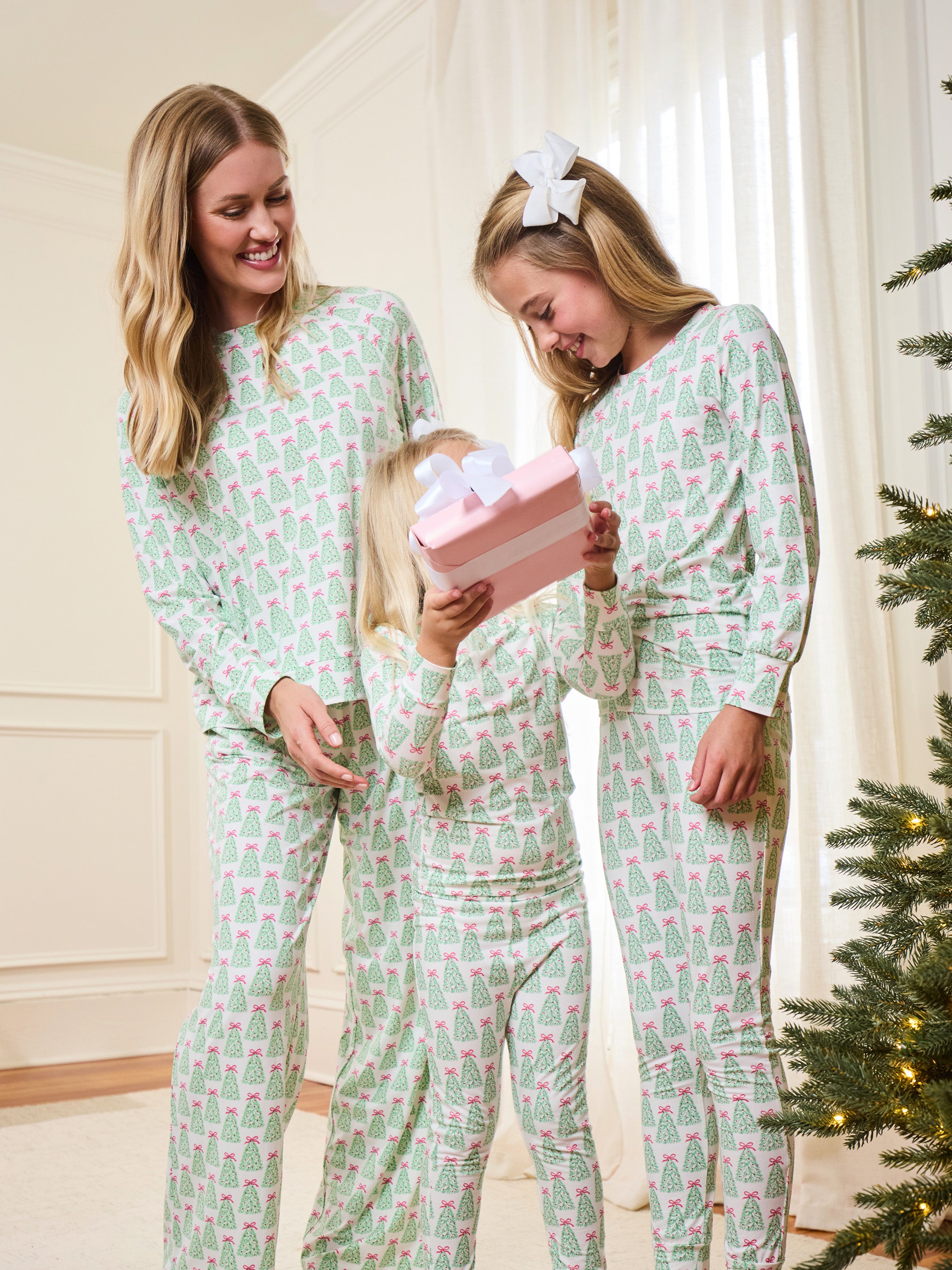 Three women of different ages wearing matching light green and pink Christmas tree print pajamas, standing together in a brightly lit room next to a Christmas tree and holding a pink gift box.