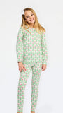 A young girl with blonde hair and a white bow in her hair is wearing a two-piece pajama set with a green and pink Christmas tree pattern, standing against a plain white background.