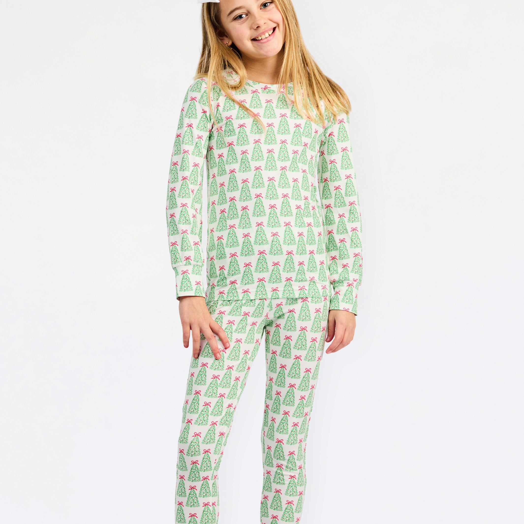 A young girl with blonde hair and a white bow in her hair is wearing a two-piece pajama set with a green and pink Christmas tree pattern, standing against a plain white background.