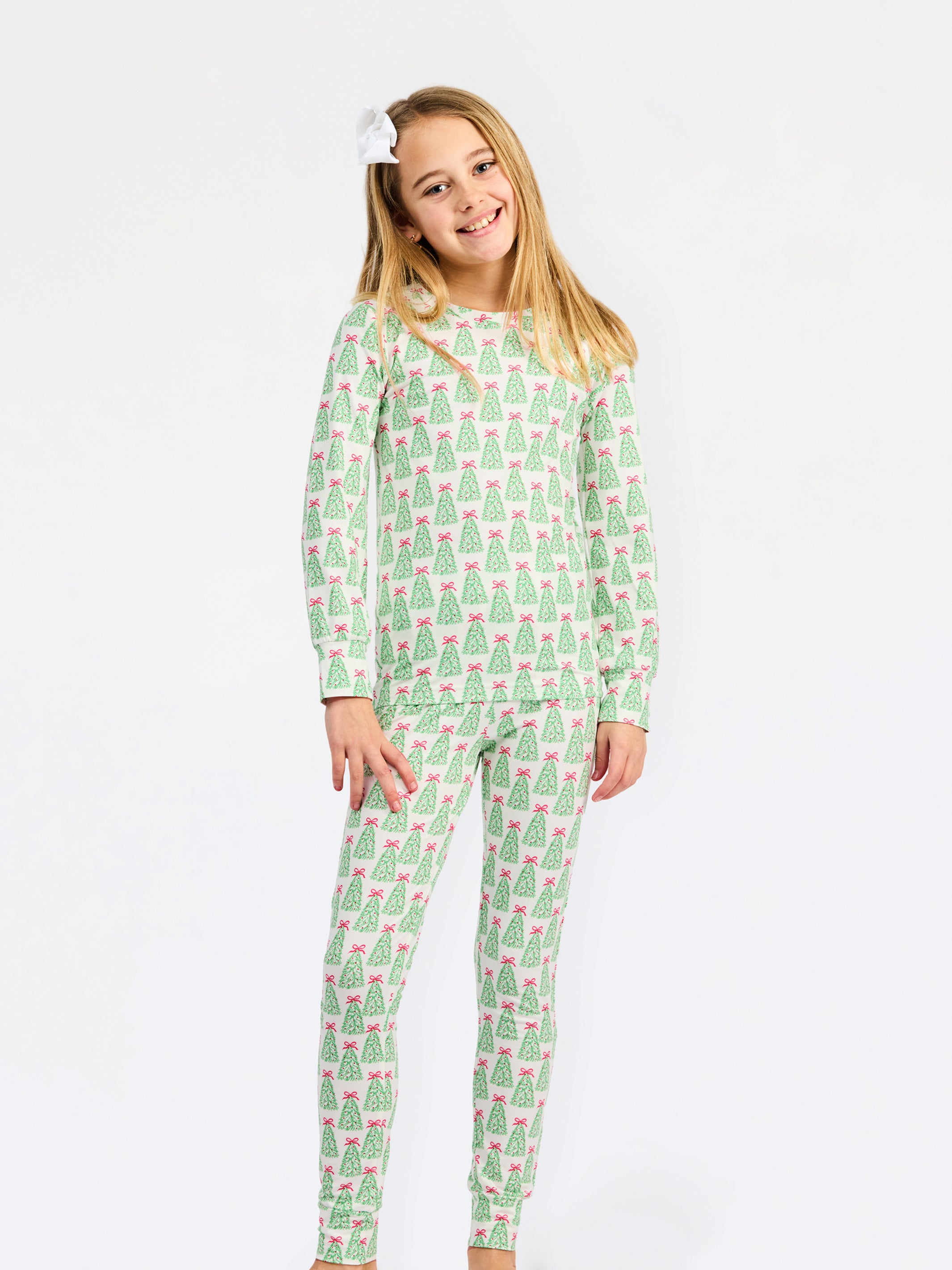 A young girl with blonde hair and a white bow in her hair is wearing a two-piece pajama set with a green and pink Christmas tree pattern, standing against a plain white background.