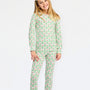 A young girl with blonde hair and a white bow in her hair is wearing a two-piece pajama set with a green and pink Christmas tree pattern, standing against a plain white background.