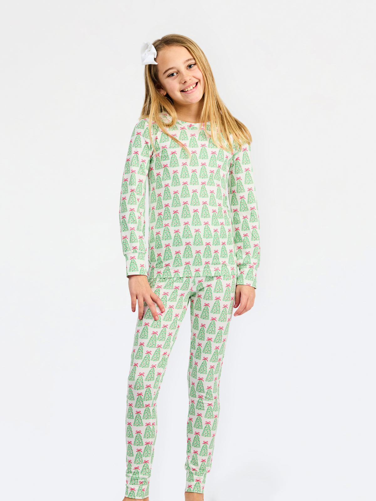 A young girl with blonde hair and a white bow in her hair is wearing a two-piece pajama set with a green and pink Christmas tree pattern, standing against a plain white background.