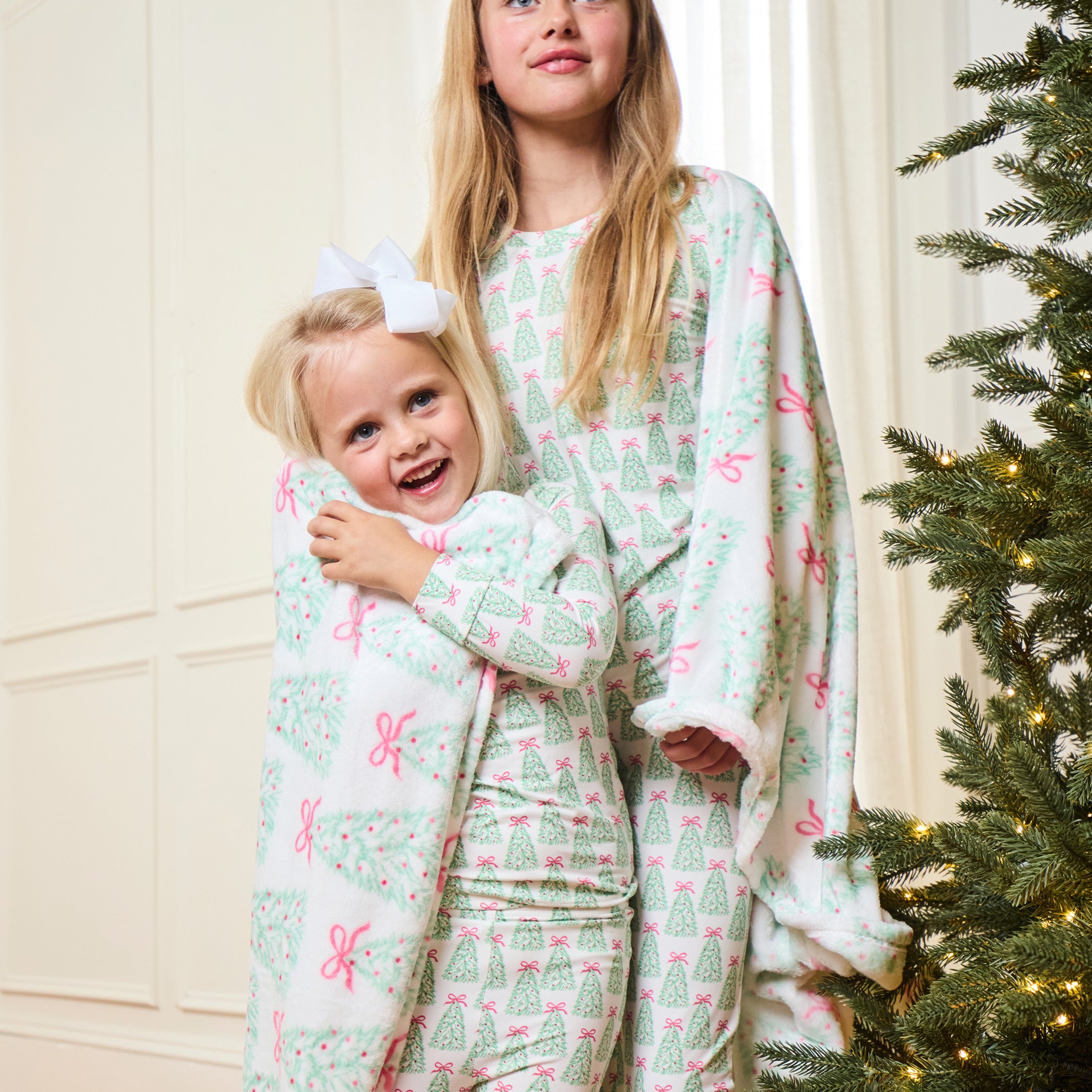 Two young girls wearing matching Christmas-themed pajamas with a green tree and pink bow pattern, standing in a warmly lit room next to a decorated Christmas tree.