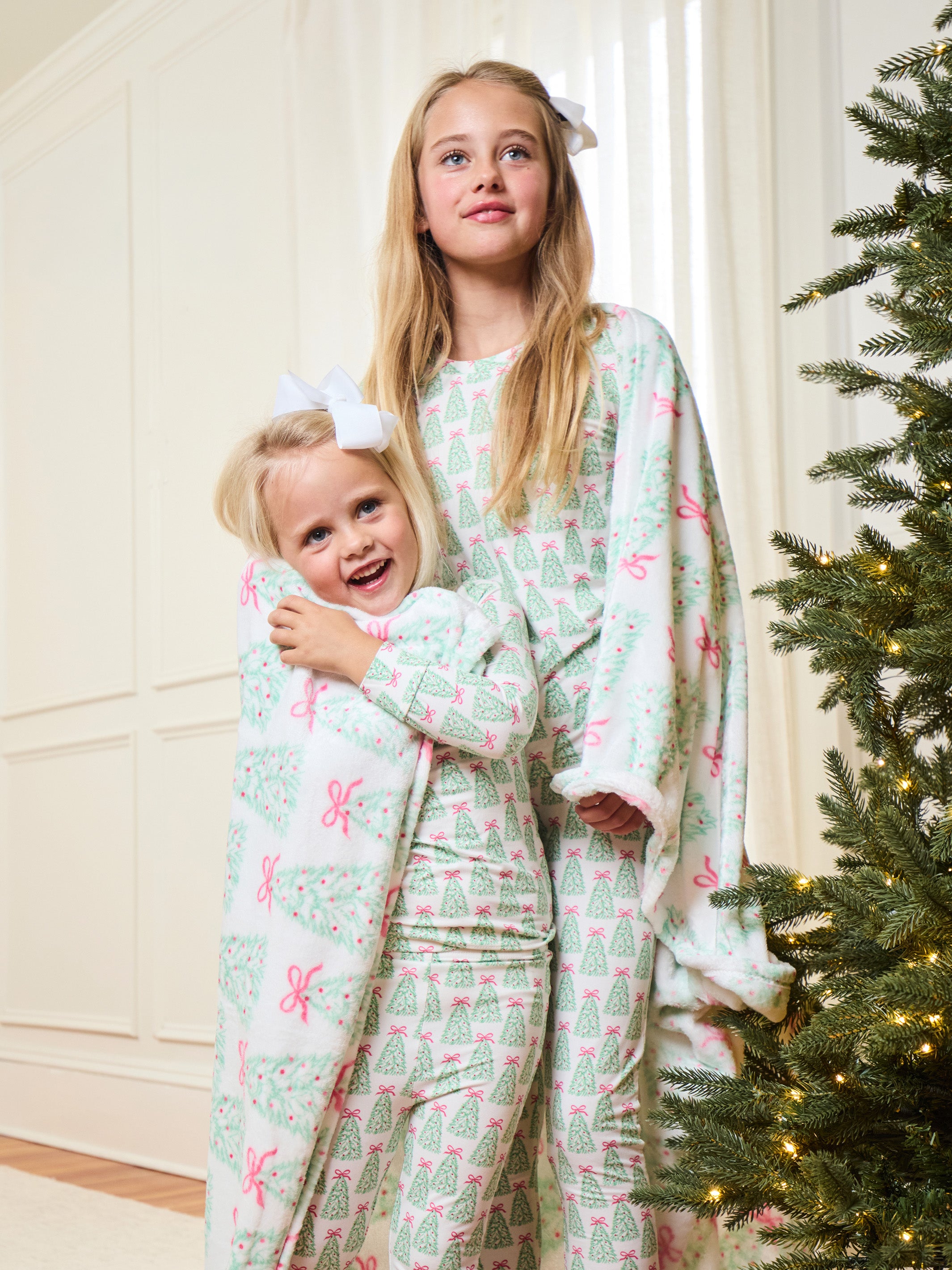 Two young girls wearing matching Christmas-themed pajamas with a green tree and pink bow pattern, standing in a warmly lit room next to a decorated Christmas tree.