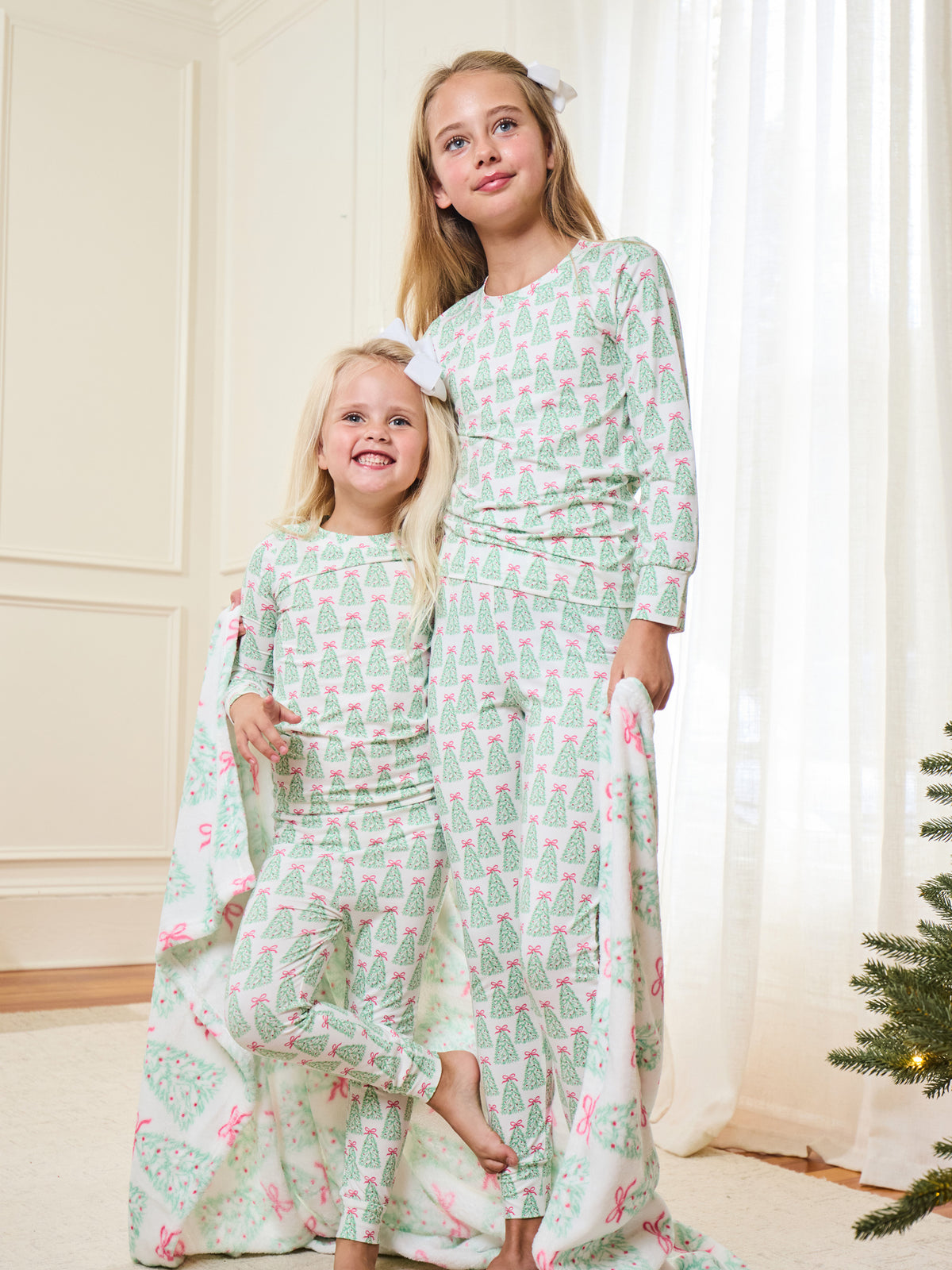 Two young girls wearing matching long-sleeve pajama sets with a mint green and pink Christmas tree pattern, standing indoors in a bright room with white curtains and a small Christmas tree visible at the edge.