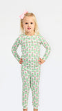 A young girl with blonde hair and a pink bow stands against a white studio background wearing a long-sleeve pajama set with an all-over green Christmas tree pattern and pink bows.