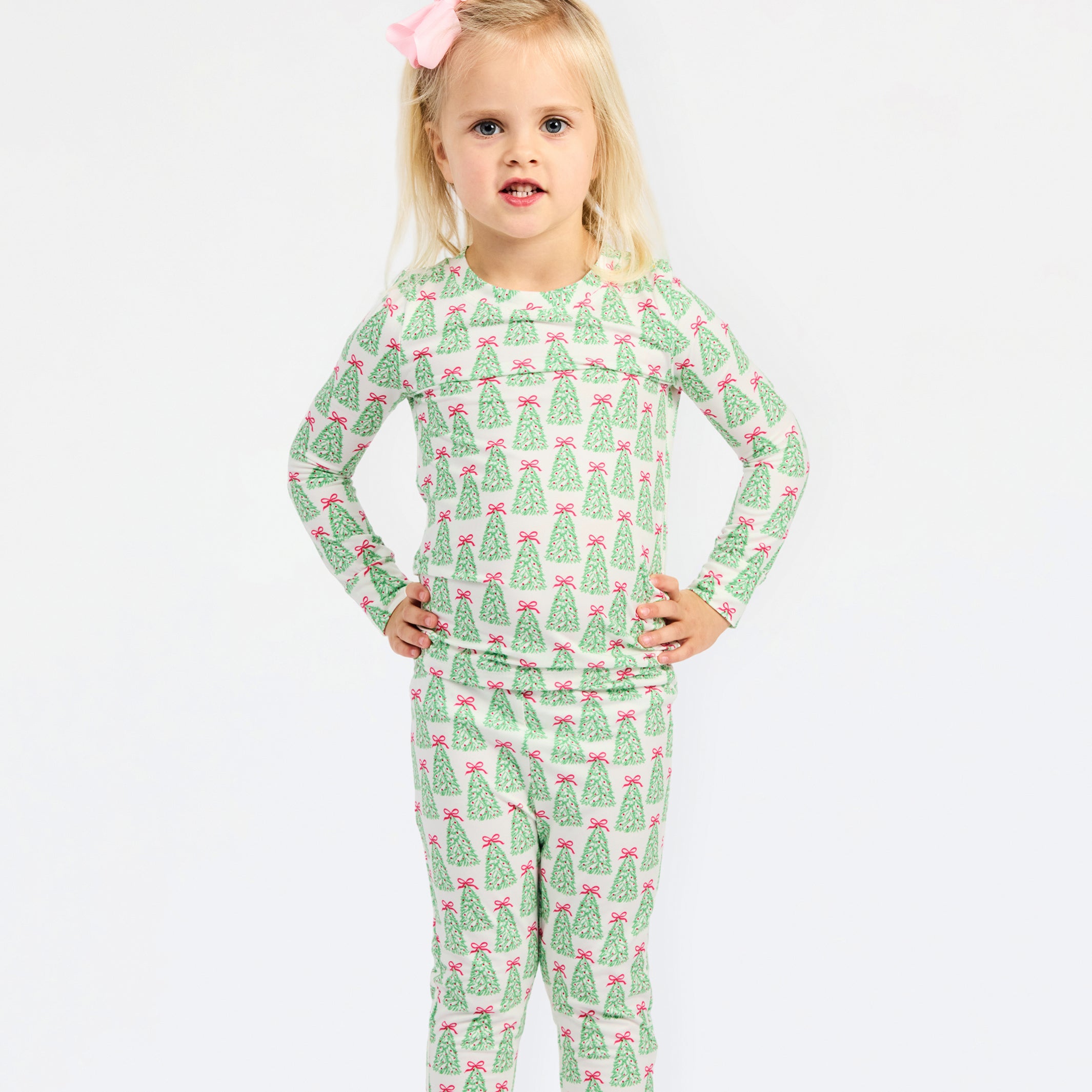 A young girl with blonde hair and a pink bow stands against a white studio background wearing a long-sleeve pajama set with an all-over green Christmas tree pattern and pink bows.
