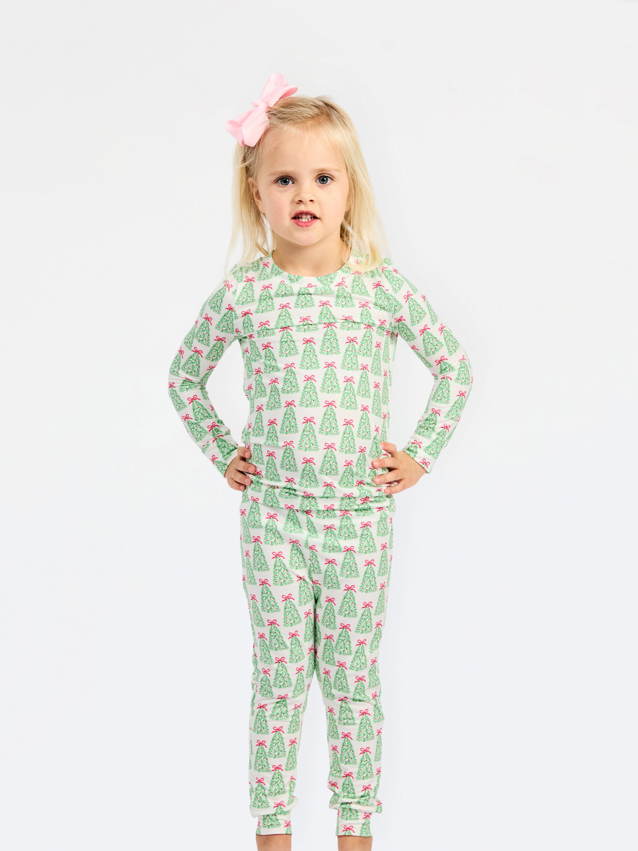 A young girl with blonde hair and a pink bow stands against a white studio background wearing a long-sleeve pajama set with an all-over green Christmas tree pattern and pink bows.