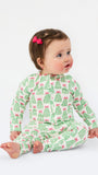 A baby sitting on a white studio background wearing a green and pink Christmas tree print long-sleeve romper. The baby has brown hair with a pink bow and is looking to the side.