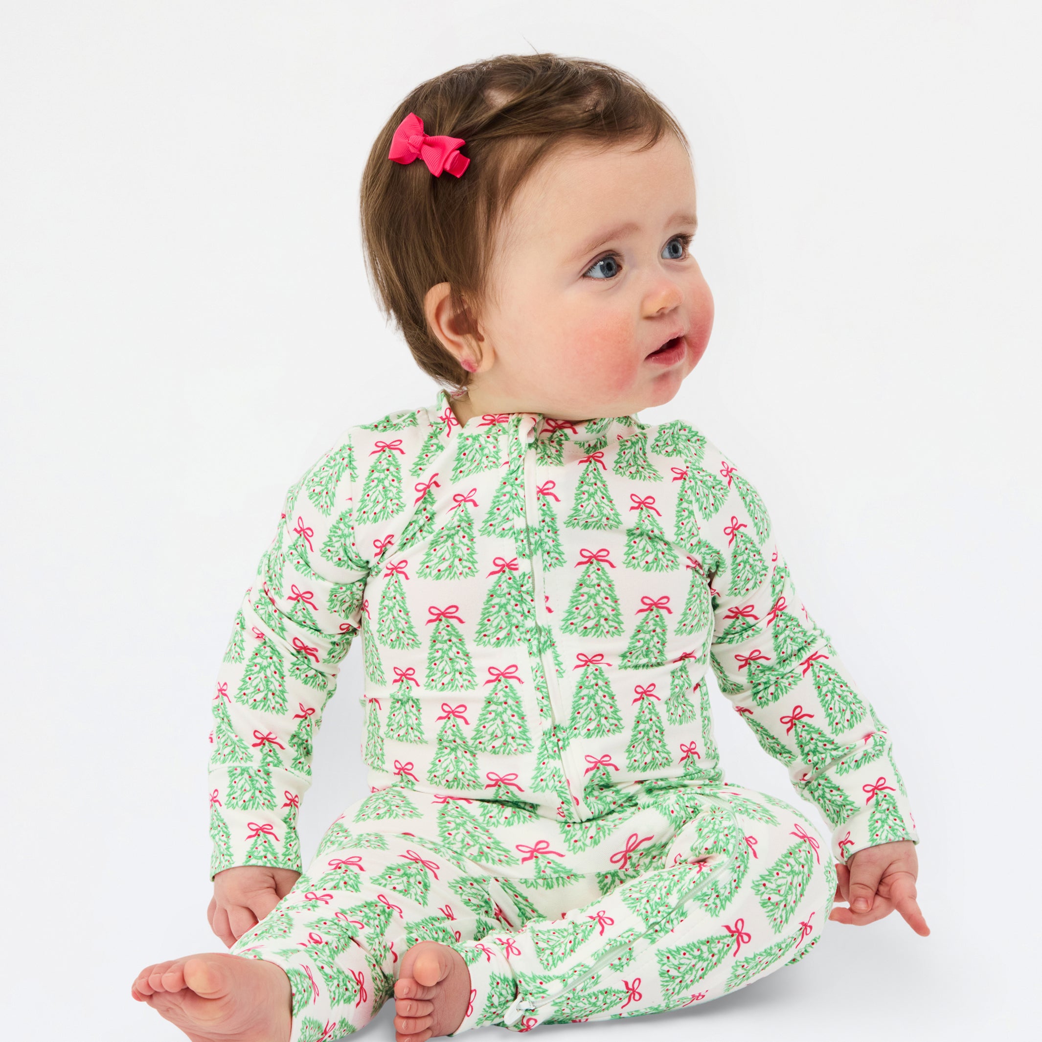 A baby sitting on a white studio background wearing a green and pink Christmas tree print long-sleeve romper. The baby has brown hair with a pink bow and is looking to the side.