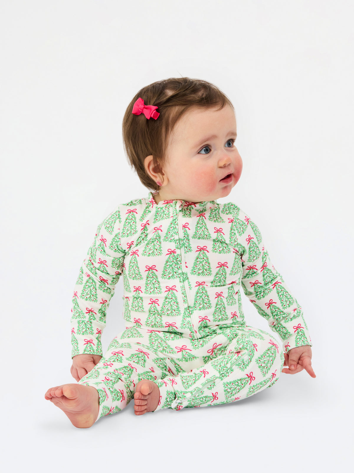 A baby sitting on a white studio background wearing a green and pink Christmas tree print long-sleeve romper. The baby has brown hair with a pink bow and is looking to the side.