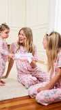 Three women wearing matching pink pajamas with a green and white holiday tree print are gathered in a brightly lit bedroom. One woman stands on the left, one sits kneeling in the center holding a white gift box with a bow, and one sits on the right.