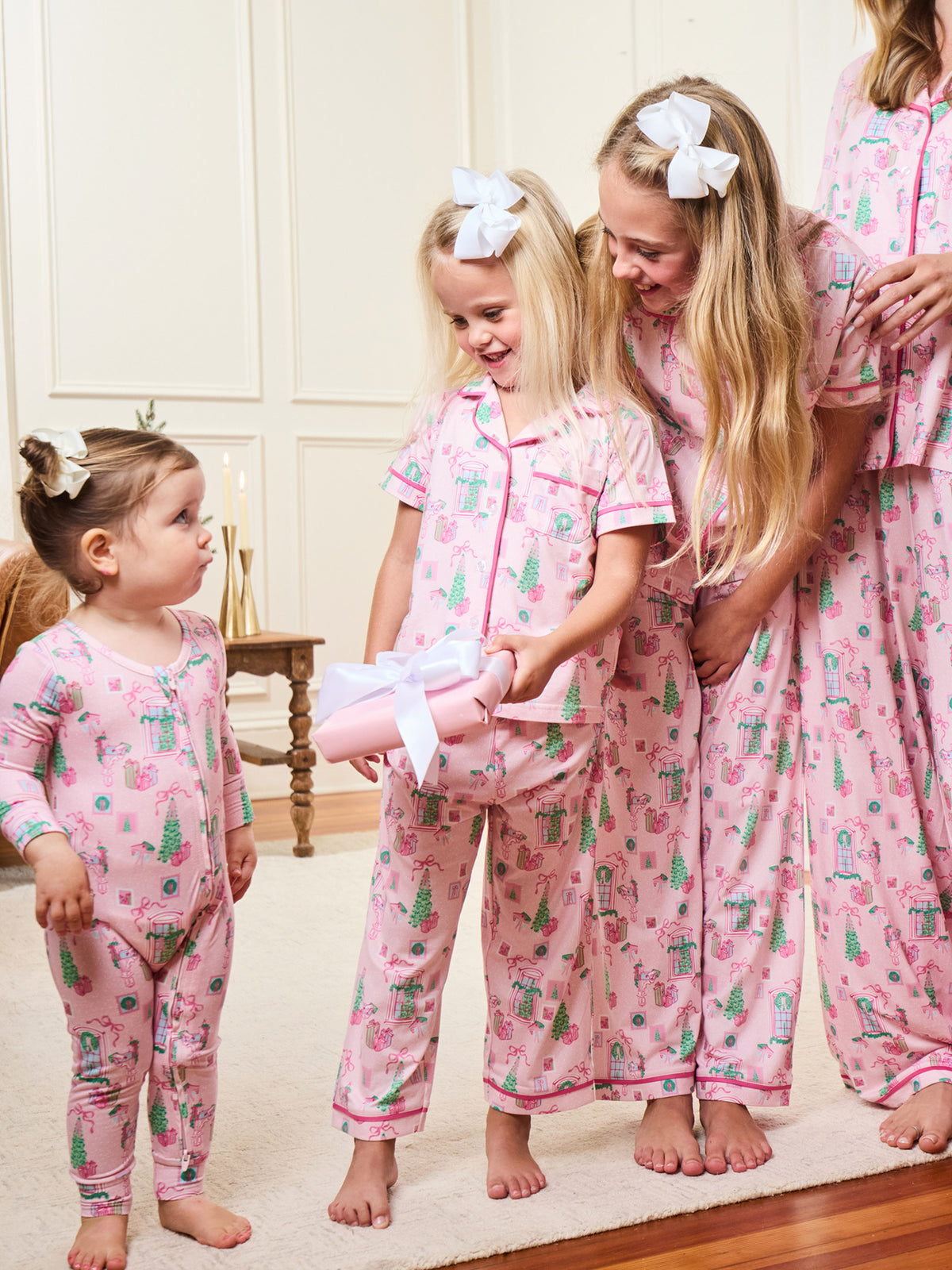 Four women wearing matching pink Christmas-themed pajamas with a tree print, standing together in a warmly lit room. The pajamas feature a repeating pattern of green trees, red ornaments, and white bows.