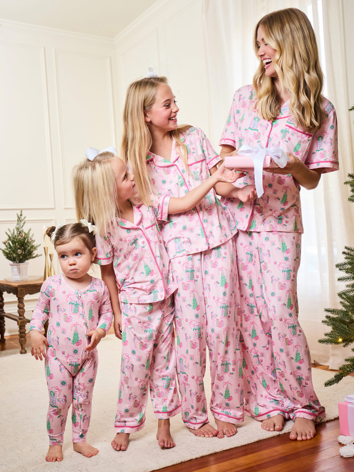 Four women wearing matching pink cactus-print pajamas pose together in a bright room, with one woman holding a wrapped gift box.