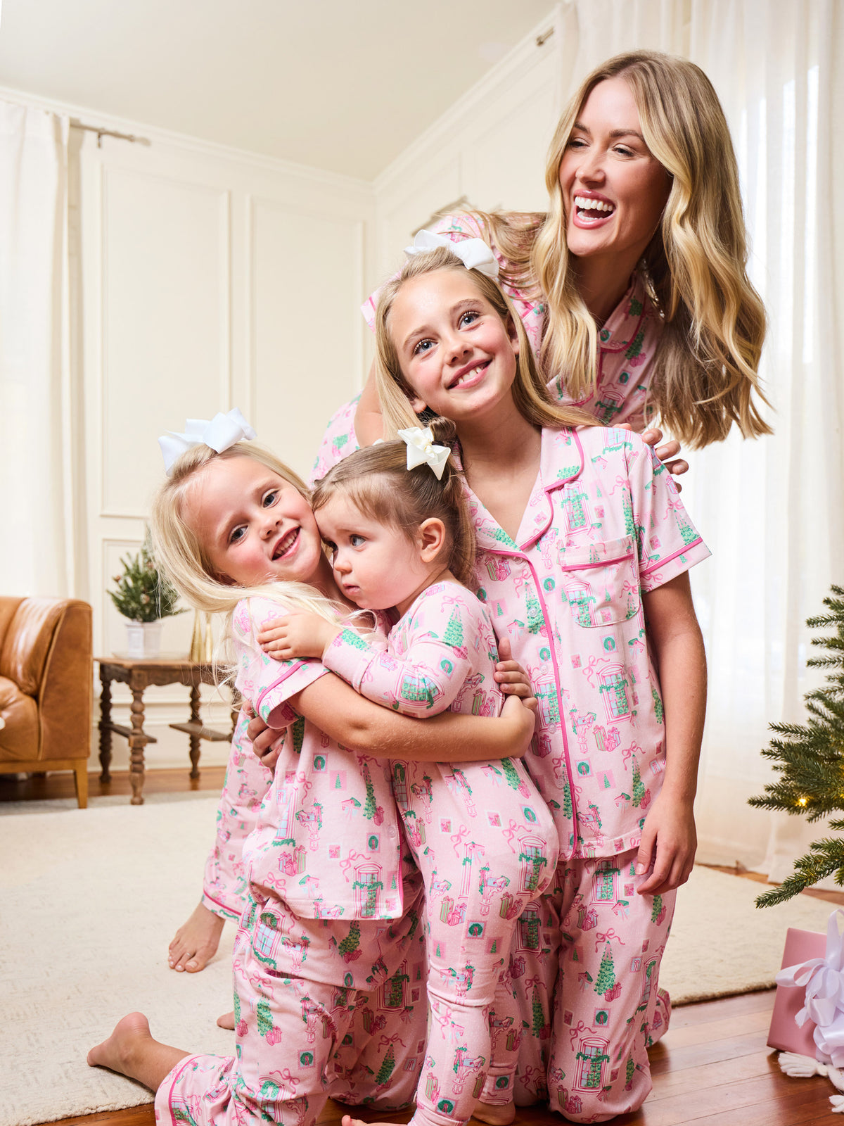 A woman and three young girls wearing matching pink cactus-print pajamas hugging in a cozy, warmly lit bedroom setting.