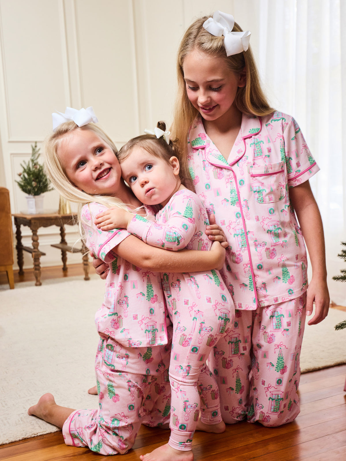 Three young girls wearing matching pink pajamas with a green and blue Christmas tree pattern, posing together in a brightly lit room.