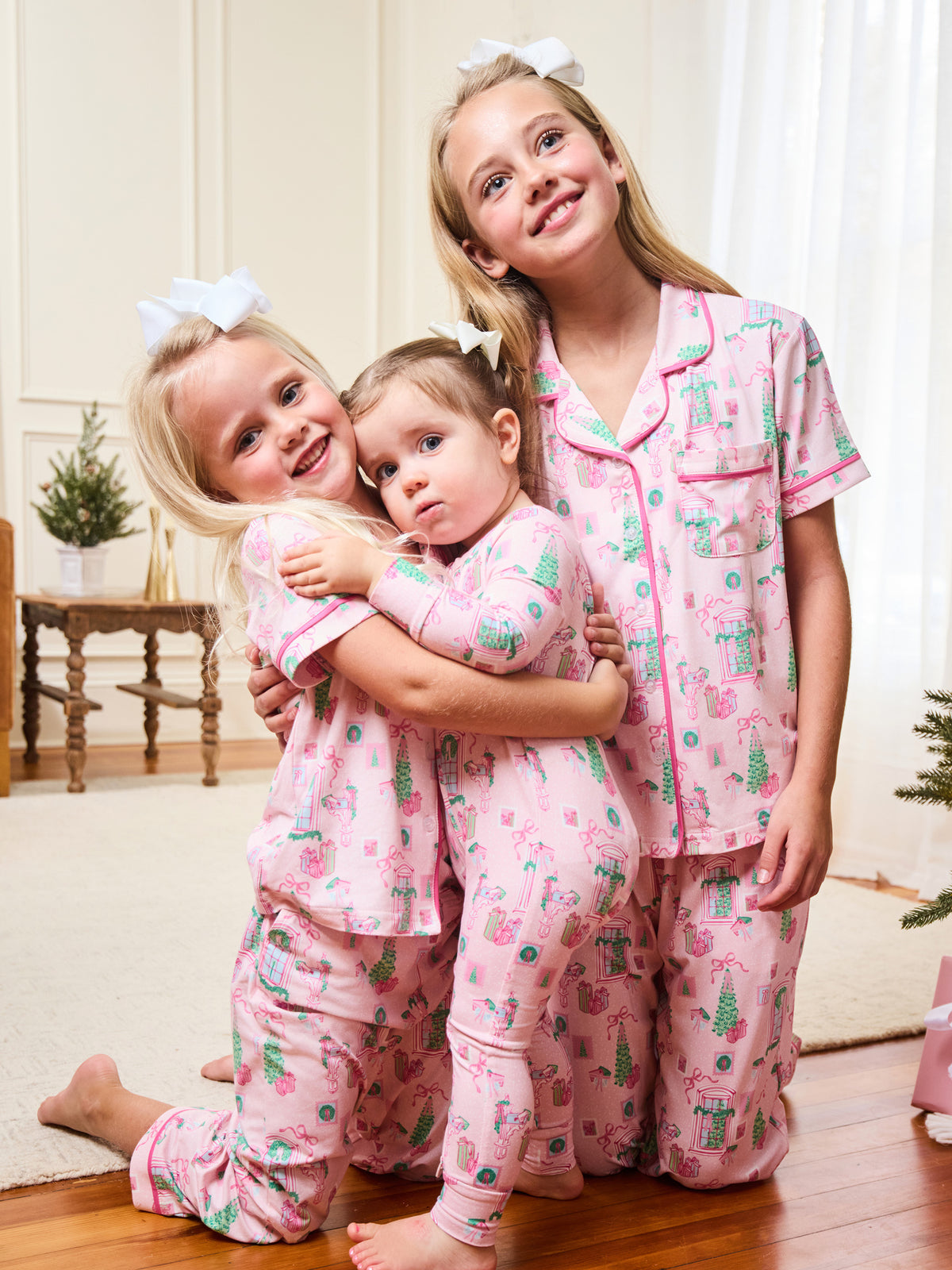 Three young girls wearing matching pink pajamas with a green and white holiday tree and candy cane print, posed together in a cozy indoor room setting.