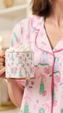 A person wearing pink pajamas with a holiday village print holds a mug topped with whipped cream in a warmly lit indoor setting.