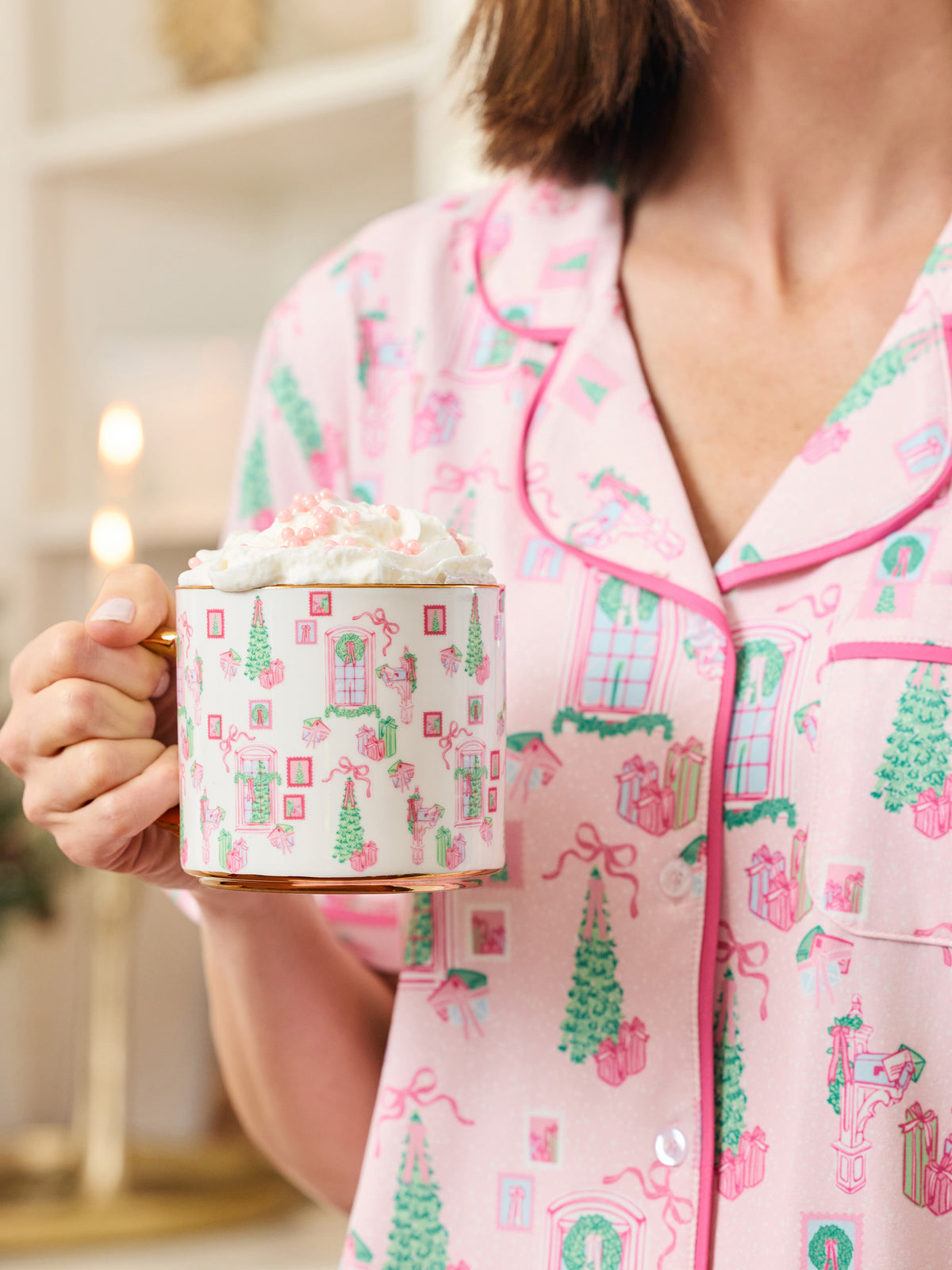 A person wearing pink pajamas with a holiday village print holds a mug topped with whipped cream in a warmly lit indoor setting.