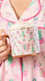 A person in pink holiday pajamas holds a white ceramic mug with a gold handle and base, decorated with pink and green Christmas gift and tree motifs. The mug is filled with whipped cream and topped with pink sprinkles.