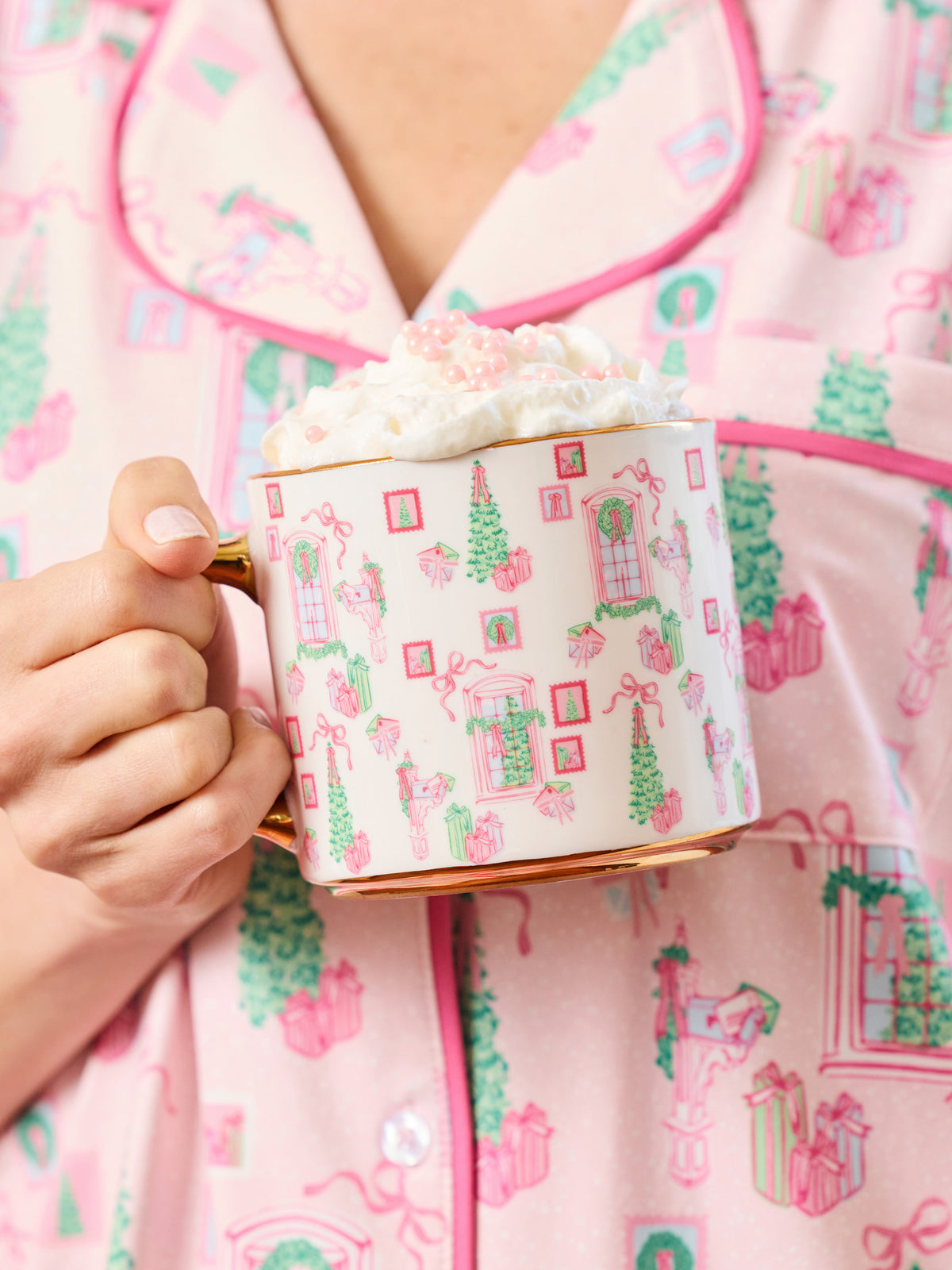 A person in pink holiday pajamas holds a white ceramic mug with a gold handle and base, decorated with pink and green Christmas gift and tree motifs. The mug is filled with whipped cream and topped with pink sprinkles.