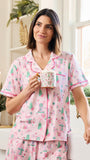 A woman wearing a pink short-sleeve pajama set with a green and white holiday print, holding a mug in a bright, modern bedroom setting.