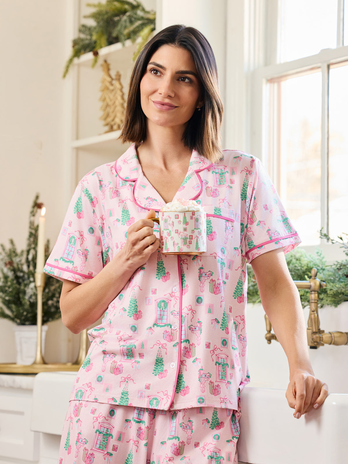 A woman wearing a pink short-sleeve pajama set with a green and white holiday print, holding a mug in a bright, modern bedroom setting.