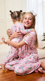 Three children wearing matching pink Christmas-themed pajamas hugging on a wooden floor in a living room with a Christmas tree and wrapped presents.