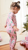 A toddler stands barefoot in a living room wearing a pink long-sleeve pajama set with a Christmas tree and snowman print. A woman in matching pajamas kneels on the left, and a Christmas tree with wrapped presents is visible on the right.