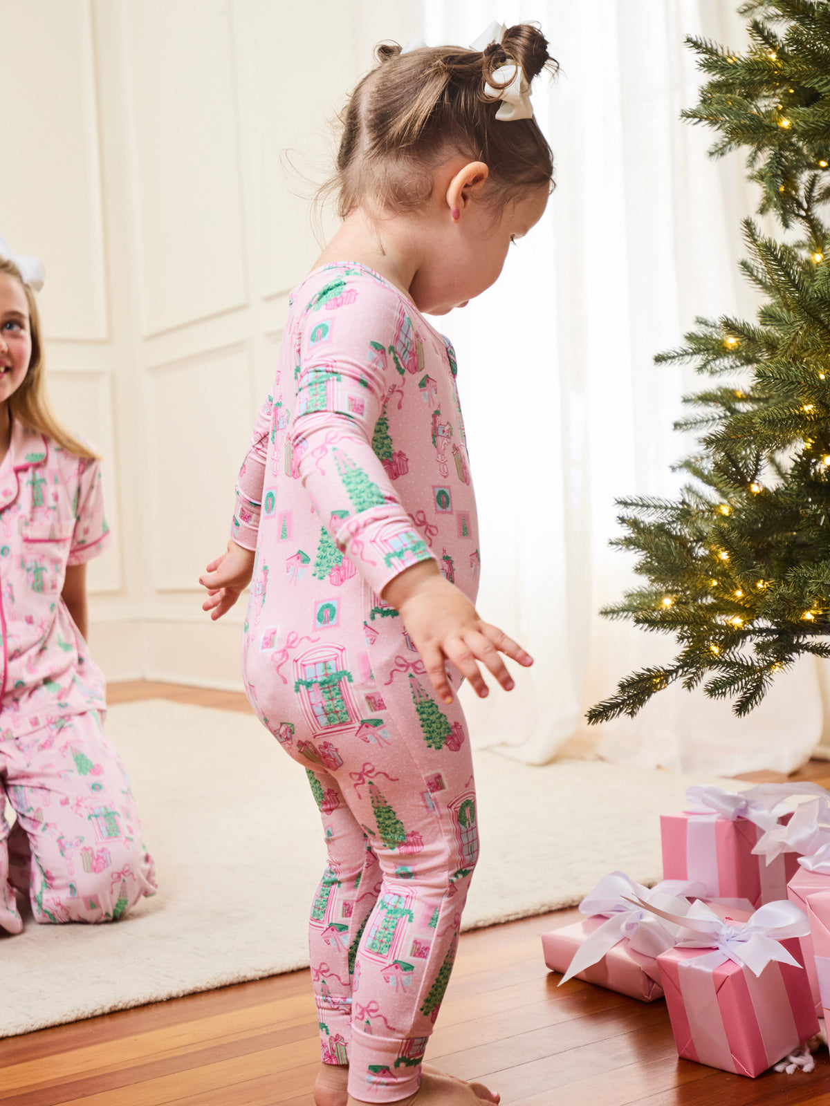 A toddler stands barefoot in a living room wearing a pink long-sleeve pajama set with a Christmas tree and snowman print. A woman in matching pajamas kneels on the left, and a Christmas tree with wrapped presents is visible on the right.