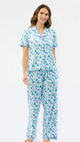 A woman modeling a two-piece pajama set with a white base and an all-over green and blue Christmas tree pattern, set against a plain white studio background.