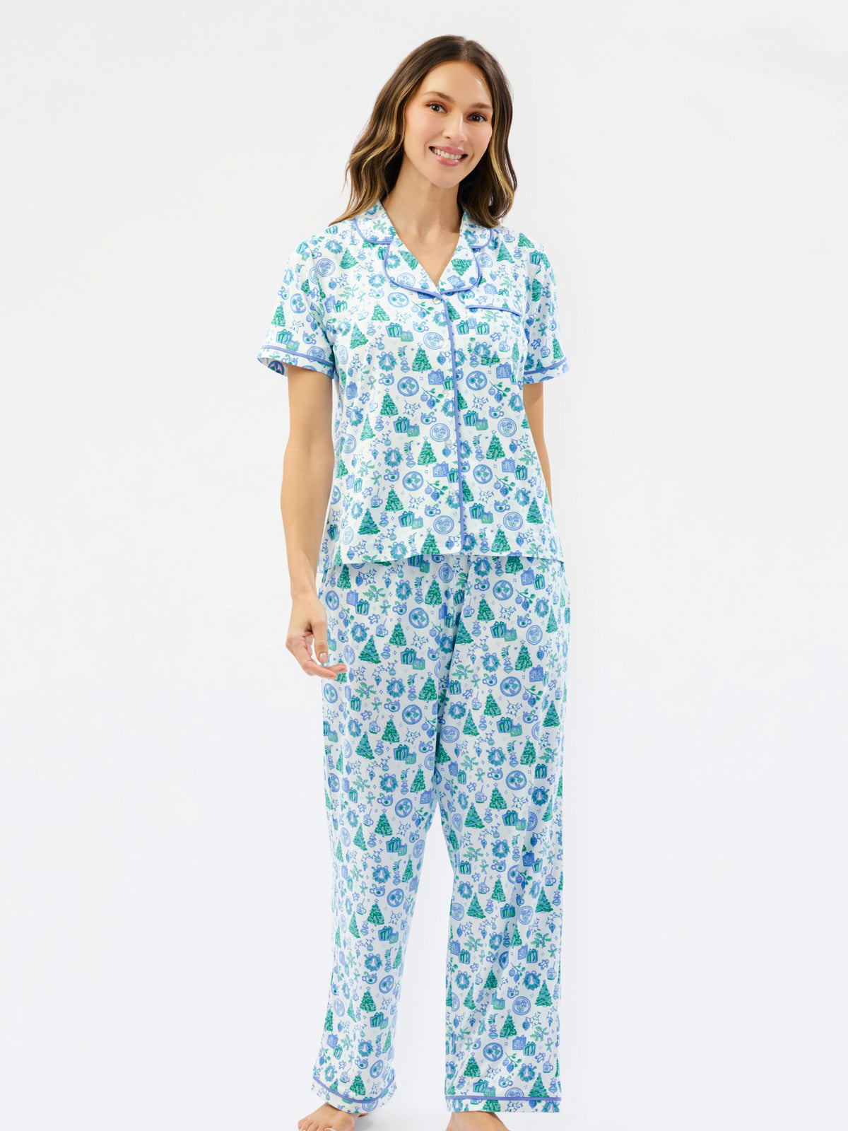 A woman modeling a two-piece pajama set with a white base and an all-over green and blue Christmas tree pattern, set against a plain white studio background.