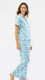 A woman modeling a two-piece pajama set with a short-sleeve button-up top and matching pants, featuring a white base with a repeating pattern of blue and green pine trees and snowflakes, set against a plain white studio background.