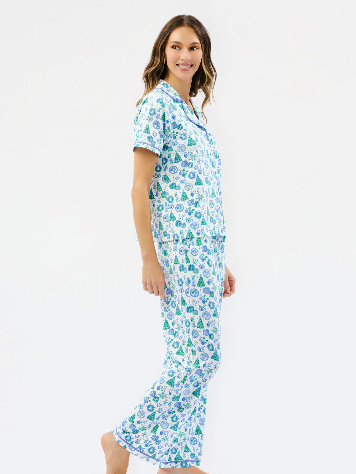 A woman modeling a two-piece pajama set with a short-sleeve button-up top and matching pants, featuring a white base with a repeating pattern of blue and green pine trees and snowflakes, set against a plain white studio background.