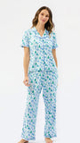 A woman modeling a matching short-sleeve button-up pajama top and wide-leg pajama pants set, featuring a light blue and white background with a green pine tree and snowflake pattern, against a plain white studio background.