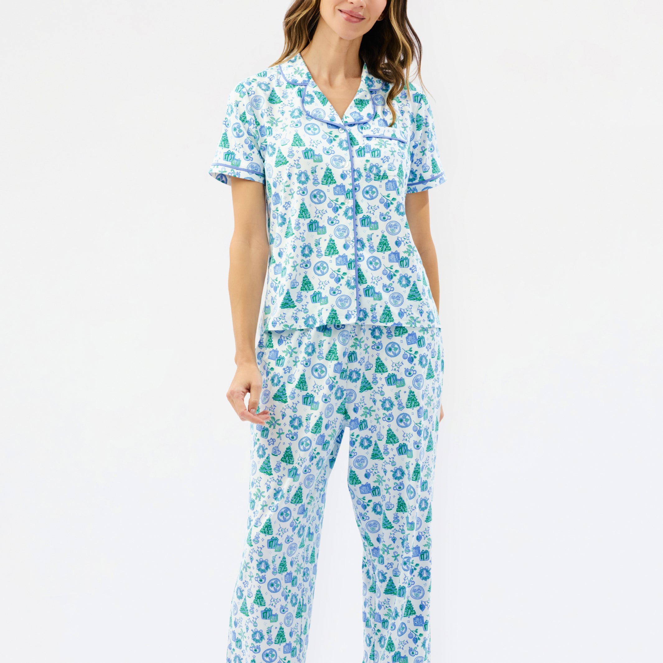 A woman modeling a matching short-sleeve button-up pajama top and wide-leg pajama pants set, featuring a light blue and white background with a green pine tree and snowflake pattern, against a plain white studio background.