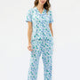 A woman modeling a matching short-sleeve button-up pajama top and wide-leg pajama pants set, featuring a light blue and white background with a green pine tree and snowflake pattern, against a plain white studio background.