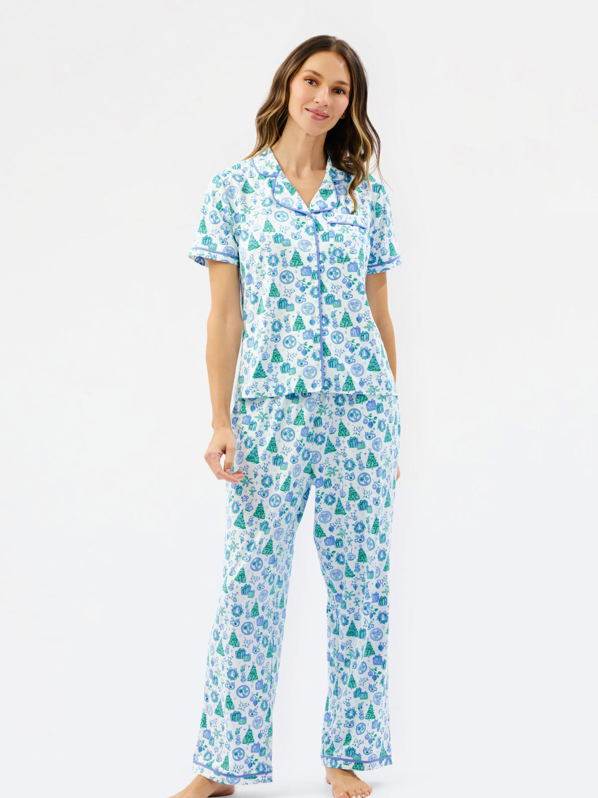 A woman modeling a matching short-sleeve button-up pajama top and wide-leg pajama pants set, featuring a light blue and white background with a green pine tree and snowflake pattern, against a plain white studio background.