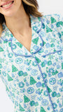 A woman modeling a matching short-sleeve button-up pajama top and wide-leg pajama pants set, featuring a light blue and white background with a green pine tree and snowflake pattern, against a plain white studio background.
