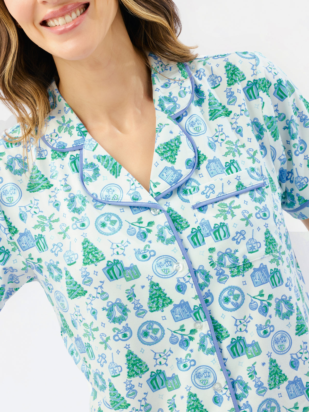 A woman modeling a matching short-sleeve button-up pajama top and wide-leg pajama pants set, featuring a light blue and white background with a green pine tree and snowflake pattern, against a plain white studio background.