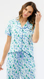 A woman modeling a matching short-sleeve button-front pajama top and drawstring pajama pants set, both printed with a blue and green holiday tree and snowflake pattern on a white background, against a plain white studio backdrop.