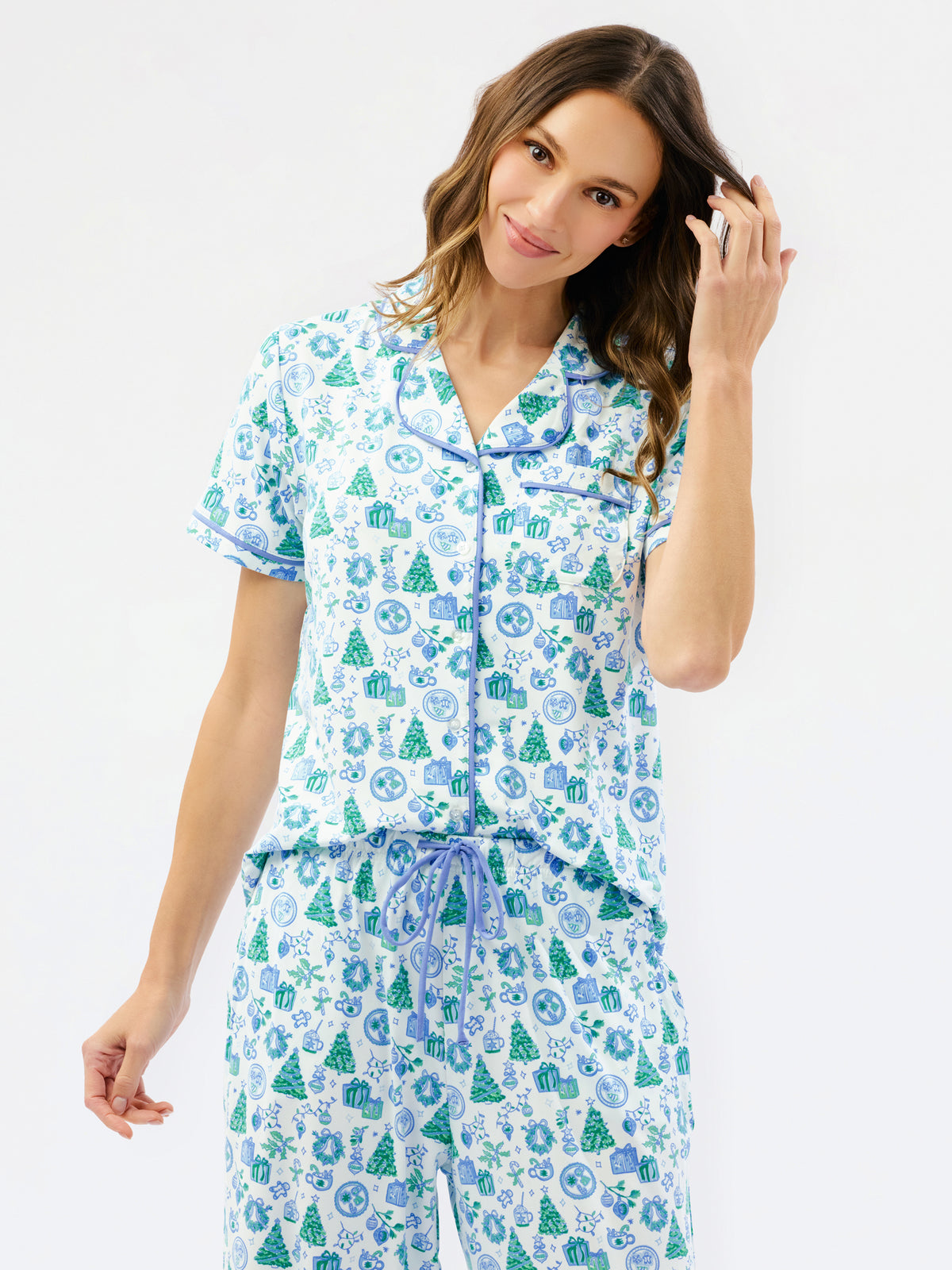A woman modeling a matching short-sleeve button-front pajama top and drawstring pajama pants set, both printed with a blue and green holiday tree and snowflake pattern on a white background, against a plain white studio backdrop.
