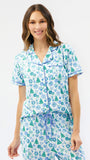 A woman modeling a matching short-sleeve pajama top and shorts set with a white base and a blue and green holiday tree pattern, set against a plain white background.